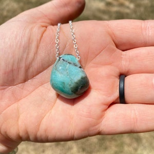 Puede incluir: Un collar de cadena de plata con un colgante de piedra preciosa verde claro y negro. El colgante tiene una forma redonda y lisa.