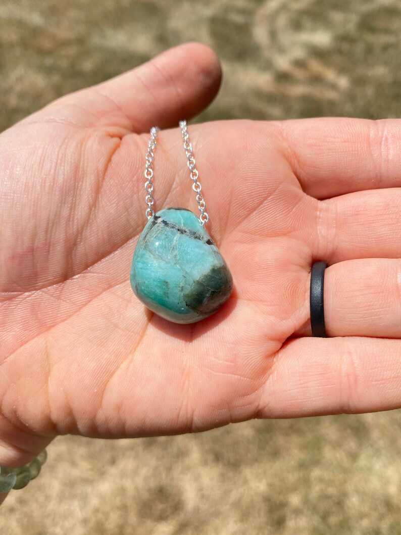 Puede incluir: Un collar de cadena de plata con un colgante de piedra preciosa verde claro y negro. El colgante tiene una forma redonda y lisa.