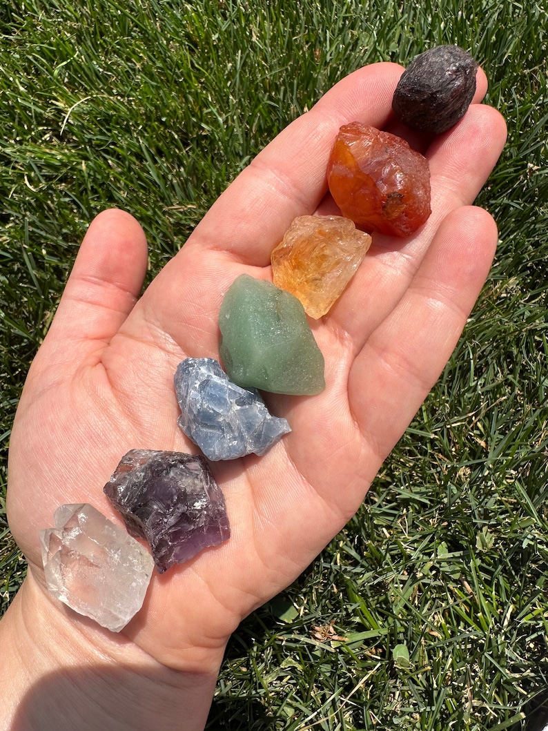 May include: A hand holding seven tumbled chakra stones in a rainbow colour order. The stones are clear quartz, amethyst, blue lace agate, green aventurine, citrine, carnelian, and black obsidian.