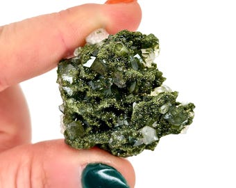 May include: A close-up of a green mineral specimen with clear, crystalline structures. The mineral has a mossy, textured appearance with a mix of dark green and clear crystal formations. The background is white.