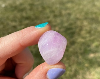 May include: A pale pink, smooth, heart-shaped crystal with white streaks. The crystal is being held by a hand with painted fingernails.