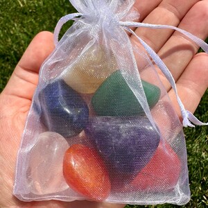 May include: A white organza bag filled with seven tumbled chakra stones. The stones are clear, yellow, green, blue, purple, red, and orange.
