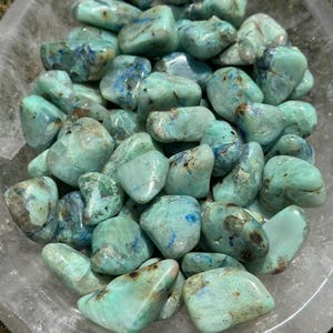 May include: A bowl of tumbled green and blue chrysocolla stones. The stones are smooth and polished, with a variety of shapes and sizes.