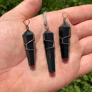 Pode incluir: Tr&ecirc;s pingentes de cristal de obsidiana preta envoltos em arame, cada um com um comprimento diferente. Os pingentes s&atilde;o exibidos em uma m&atilde;o.