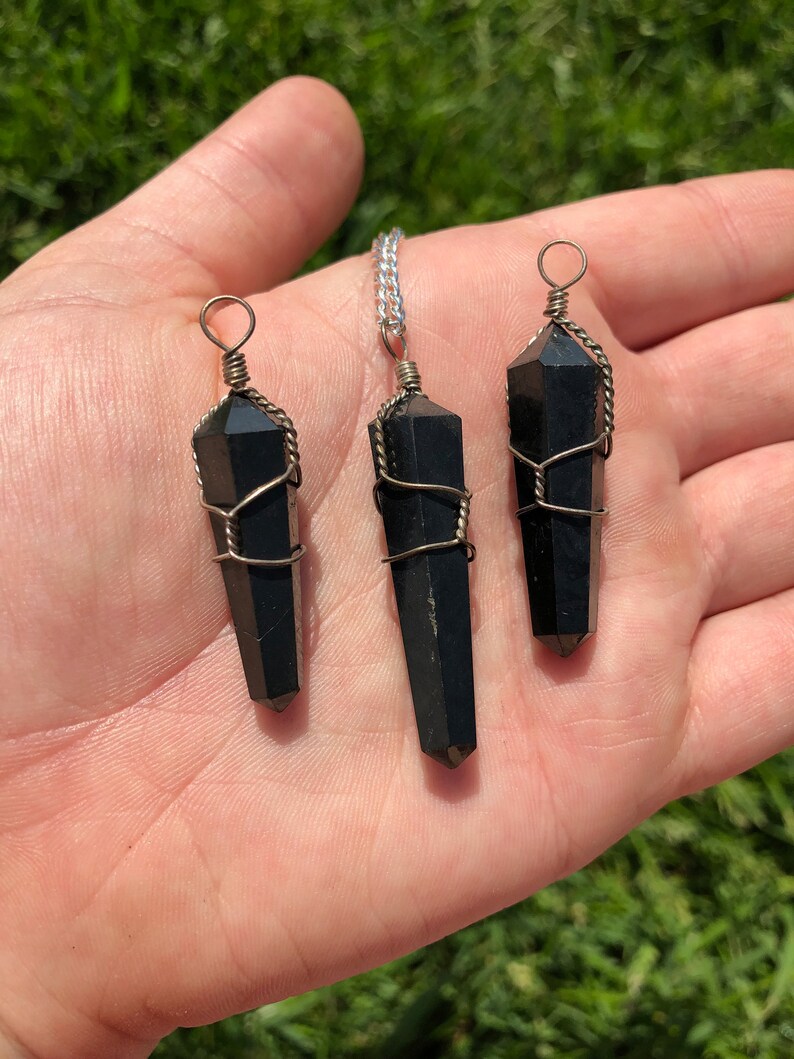 Pode incluir: Tr&ecirc;s pingentes de cristal de obsidiana preta envoltos em arame, cada um com um comprimento diferente. Os pingentes s&atilde;o exibidos em uma m&atilde;o.