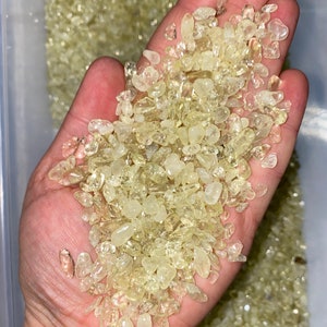 May include: A hand holding a pile of small, translucent, light yellow, and clear crystals. The crystals are smooth and have a slightly irregular shape.