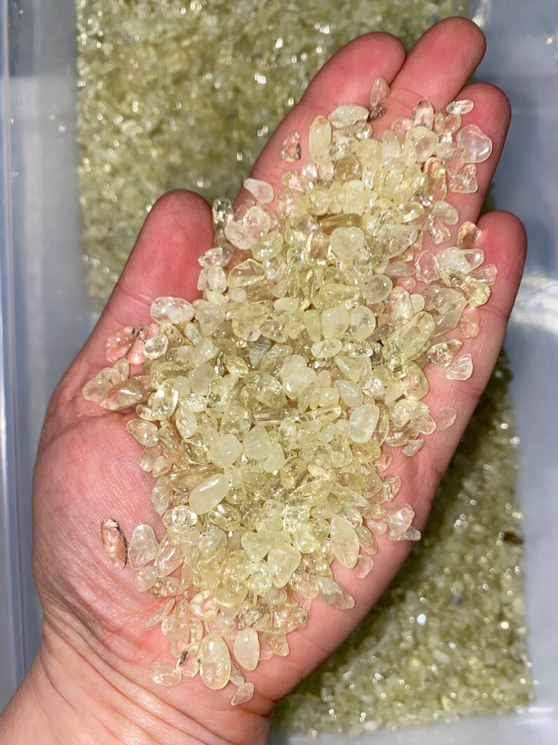 May include: A hand holding a pile of small, translucent, light yellow, and clear crystals. The crystals are smooth and have a slightly irregular shape.