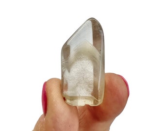 May include: A clear, elongated crystal with a light brown hue and internal cloud-like inclusions. The crystal is held between fingers against a white background, showcasing its polished surface and natural variations. The crystal's shape is slightly angled at the top.