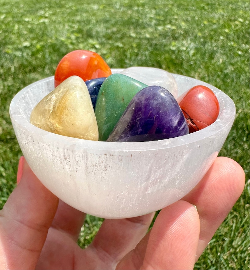 May include: A white selenite bowl filled with seven chakra stones. The stones are red, orange, yellow, green, blue, indigo, and violet.