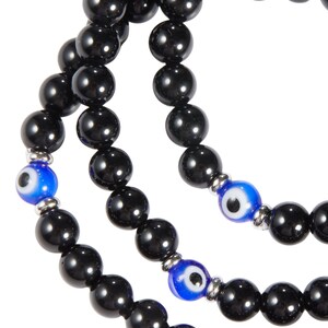 May include: Close-up of three strands of black beaded necklaces. Each strand features large, glossy black beads. Accents include blue and white evil eye beads, separated by silver-toned metal spacers. The necklaces are displayed against a white background.