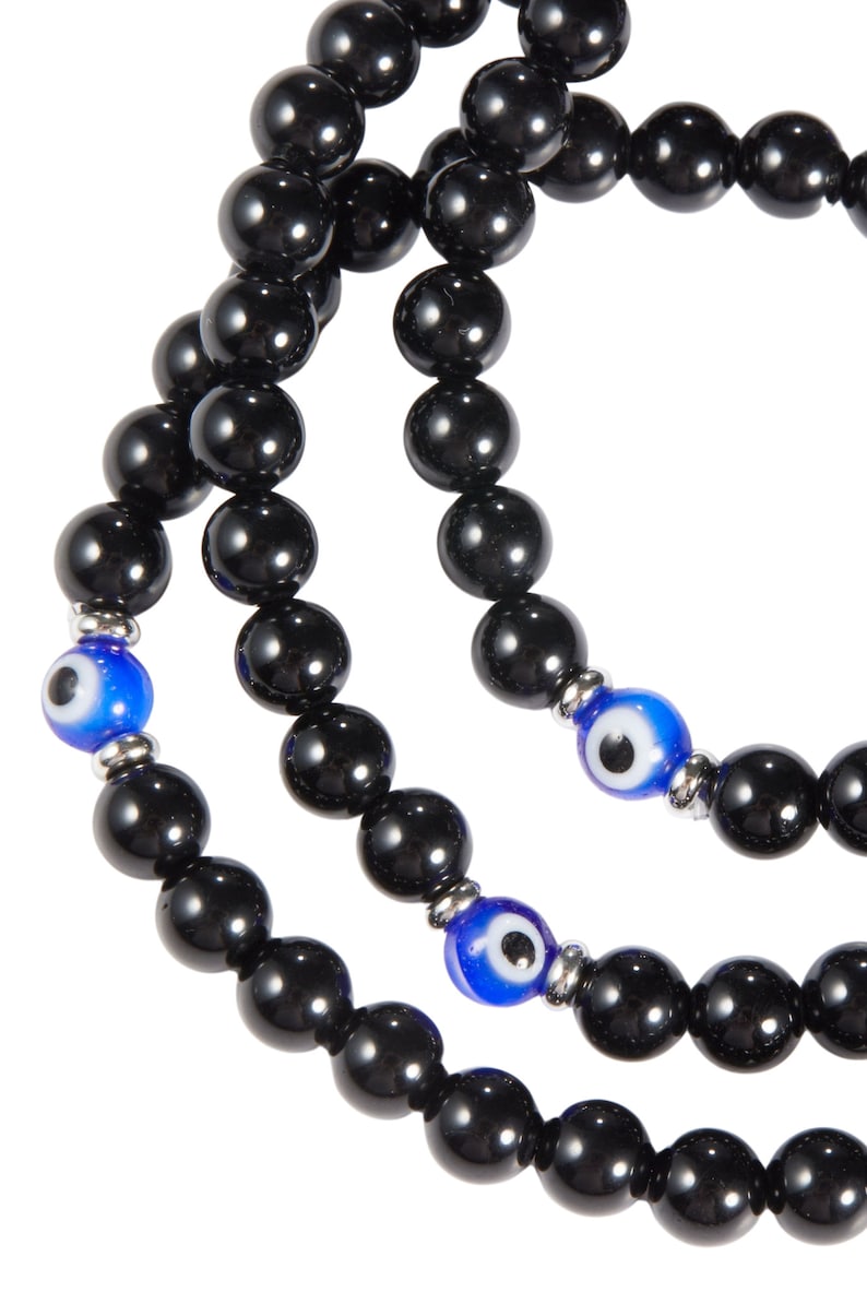 May include: Close-up of three strands of black beaded necklaces. Each strand features large, glossy black beads. Accents include blue and white evil eye beads, separated by silver-toned metal spacers. The necklaces are displayed against a white background.