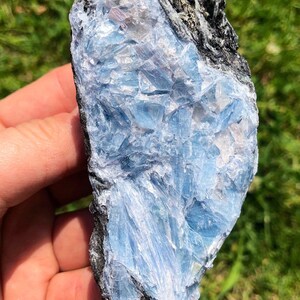 May include: A blue and gray crystal with a rough, natural texture. The crystal has a layered, striated appearance, with a darker gray band running through the center.