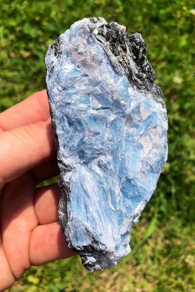 May include: A blue and gray crystal with a rough, natural texture. The crystal has a layered, striated appearance, with a darker gray band running through the center.