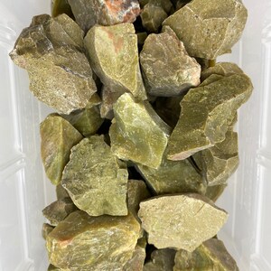 May include: A pile of rough, green, and brown serpentine stones in a clear plastic container.