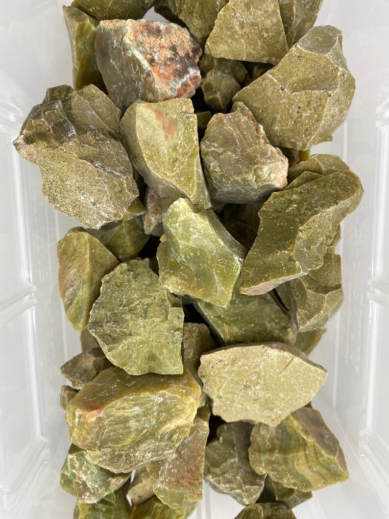 May include: A pile of rough, green, and brown serpentine stones in a clear plastic container.