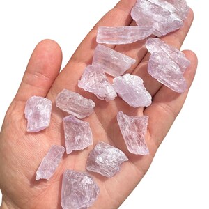 Puede incluir: Un conjunto de cristales en bruto, de color p&uacute;rpura claro, de varios tama&ntilde;os y formas, sostenidos en una mano abierta. Los cristales tienen una cualidad transl&uacute;cida, con estructuras internas visibles y una textura ligeramente rugosa. El fondo es blanco liso, resaltando los cristales.