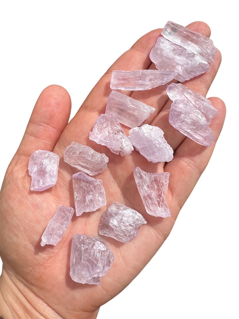Puede incluir: Un conjunto de cristales en bruto, de color p&uacute;rpura claro, de varios tama&ntilde;os y formas, sostenidos en una mano abierta. Los cristales tienen una cualidad transl&uacute;cida, con estructuras internas visibles y una textura ligeramente rugosa. El fondo es blanco liso, resaltando los cristales.