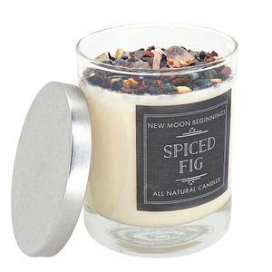 May include: A clear glass candle jar with a silver lid, filled with off-white wax topped with dark, multi-colored decorative stones and a wick. The black label reads New Moon Beginnings, Spiced Fig, and All Natural Candles in white text.