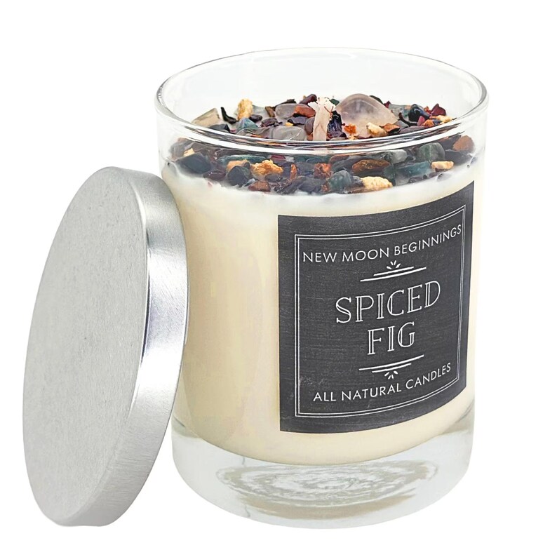 May include: A clear glass candle jar with a silver lid, filled with off-white wax topped with dark, multi-colored decorative stones and a wick. The black label reads New Moon Beginnings, Spiced Fig, and All Natural Candles in white text.
