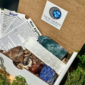 May include: An open cardboard box filled with various crystals and stones. The box contains a large green and white stone, a long white crystal, and several other colorful stones. The box also includes small informational cards and a sticker with the text "NEW MOON BEGINNINGS".