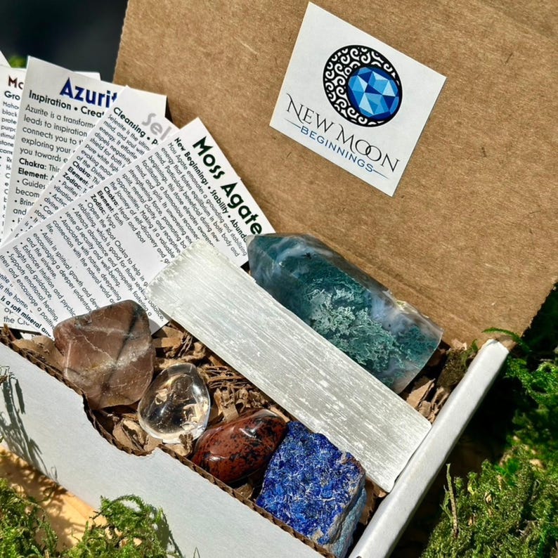 May include: An open cardboard box filled with various crystals and stones. The box contains a large green and white stone, a long white crystal, and several other colorful stones. The box also includes small informational cards and a sticker with the text "NEW MOON BEGINNINGS".