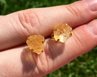 May include: A pair of gold earrings featuring raw, natural citrine stones. The stones are a light yellow color and have a rough, irregular shape.
