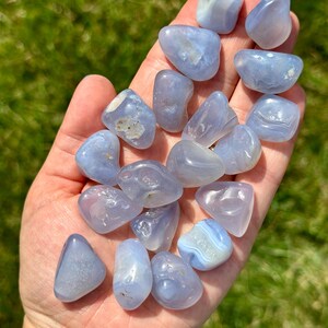 May include: A collection of tumbled blue chalcedony stones, each with a smooth, polished surface. The stones vary in shape and size, with a soft, translucent appearance. The stones are held in a hand against a backdrop of green grass.