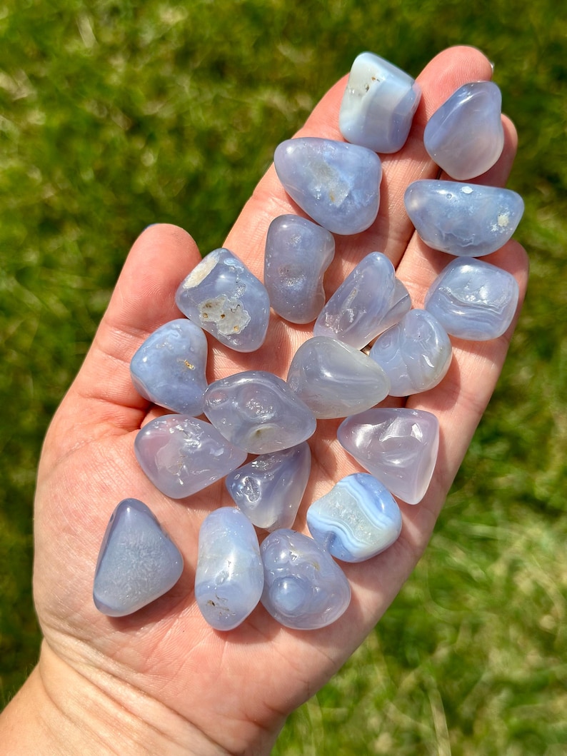 May include: A collection of tumbled blue chalcedony stones, each with a smooth, polished surface. The stones vary in shape and size, with a soft, translucent appearance. The stones are held in a hand against a backdrop of green grass.