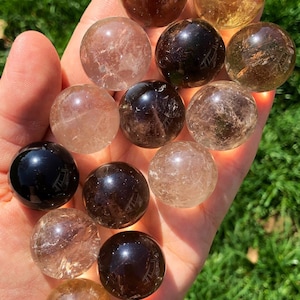 Può includere: Una collezione di 13 sfere di cristallo lisce e levigate in varie tonalità di marrone, trasparente e quarzo affumicato. Le sfere sono disposte su una mano, mostrando le loro texture e colori unici.