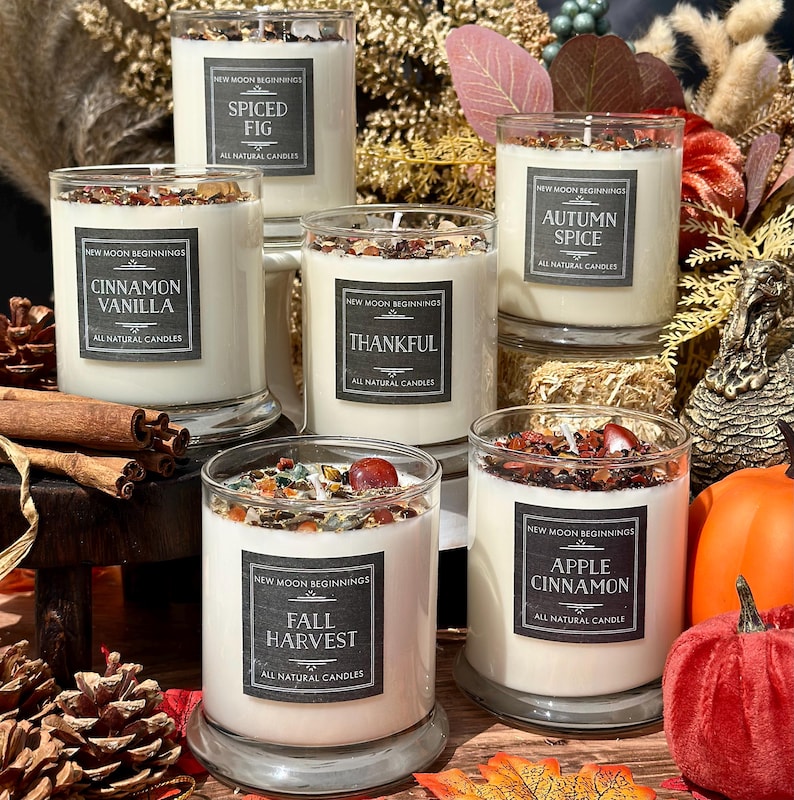 May include: A collection of five clear glass jar candles with white wax, topped with colorful crystal chips. Each candle features a black label with the text New Moon Beginnings and scent names including Spiced Fig, Cinnamon Vanilla, Thankful, Autumn Spice, Fall Harvest, and Apple Cinnamon.