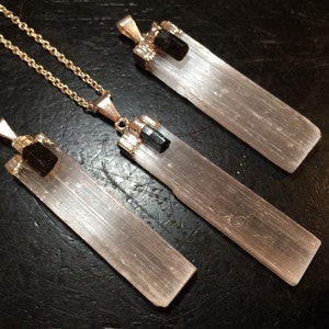 May include: Three selenite crystal pendants with black tourmaline accents. The pendants are attached to gold chains. The selenite crystals are clear and have a striated texture. The black tourmaline accents are small and round.