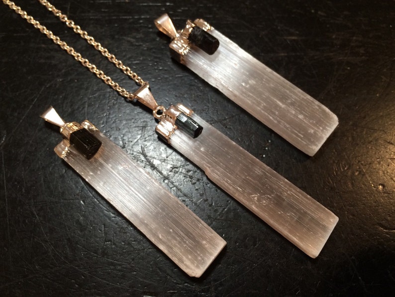 May include: Three selenite crystal pendants with black tourmaline accents. The pendants are attached to gold chains. The selenite crystals are clear and have a striated texture. The black tourmaline accents are small and round.