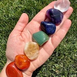 May include: Seven tumbled stones in a rainbow of colors, including clear, purple, blue, green, yellow, orange, and red. The stones are arranged in a line on a hand, with the clear stone at the top and the red stone at the bottom.
