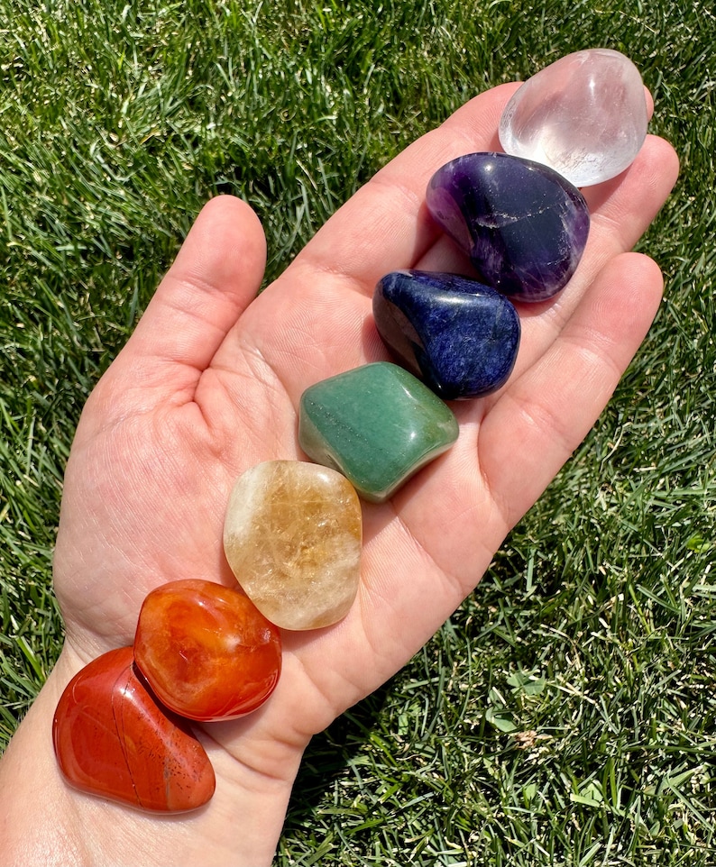 May include: Seven tumbled stones in a rainbow of colors, including clear, purple, blue, green, yellow, orange, and red. The stones are arranged in a line on a hand, with the clear stone at the top and the red stone at the bottom.