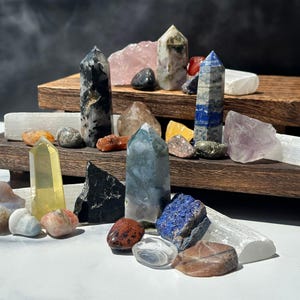 May include: A collection of various crystals and gemstones, including quartz, amethyst, and lapis lazuli, arranged on a wooden display. The crystals vary in shape, size, and color, with some polished into towers and others in their natural forms.