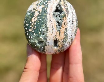 May include: A green, white and pink agate sphere with a unique pattern of dots and swirls. The sphere is smooth and polished, and it has a natural hole in the side.