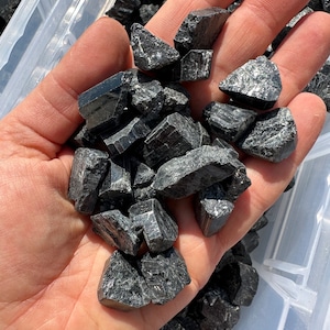 May include: A hand holding a pile of black tourmaline crystals. The crystals are rough and have a variety of shapes and sizes.