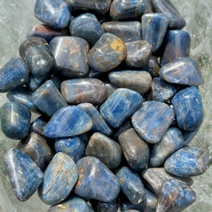 Puede incluir: Una colección de piedras de cianita azul pulidas en un cuenco de vidrio transparente. Las piedras son de varios tonos de azul y tienen un acabado liso y pulido.