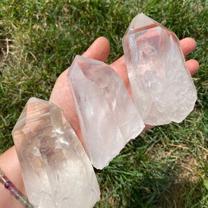 May include: Three clear quartz crystal points, also known as rock crystals, are displayed on a hand. The crystals are various sizes and shapes, with a natural, raw finish.