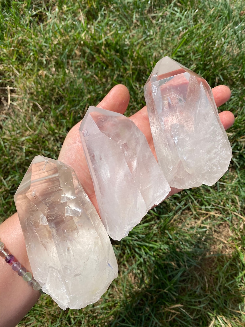May include: Three clear quartz crystal points, also known as rock crystals, are displayed on a hand. The crystals are various sizes and shapes, with a natural, raw finish.