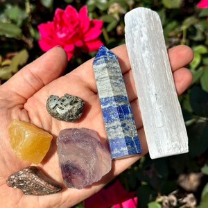 May include: A collection of crystals and gemstones in various shapes and colors. Includes a blue and gray lapis lazuli obelisk, a white selenite wand, a yellow calcite, a purple fluorite, and a small pyrite stone. The crystals are held in a hand.