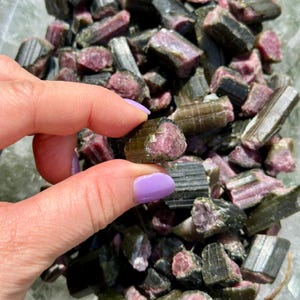 May include: A selection of raw tourmaline crystals, with a focus on a piece held in a hand. The crystals display a mix of colours, including dark green, black, and pink. The crystals have a rough, natural texture.
