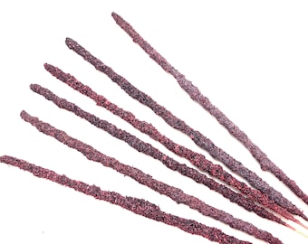 May include: Seven incense sticks with a textured, dark purple and red surface are arranged diagonally against a white background. The incense sticks have a granular appearance and are tapered at the ends.