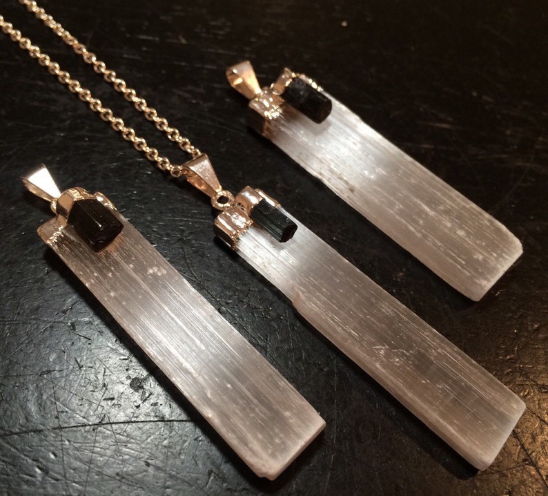 May include: Three selenite crystal pendants with black tourmaline accents. The pendants are attached to silver chains. The selenite crystals are white and translucent, while the tourmaline accents are black and opaque.
