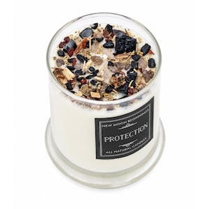 May include: A clear glass candle jar filled with white wax, topped with black stones, small brown wood pieces, and a central wick. A black rectangular label on the front reads: NEW MOON BEGINNINGS PROTECTION ALL NATURAL CANDLES.