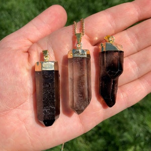 May include: Three smoky quartz crystal pendants with gold-toned caps and bails. The crystals are faceted and have a dark brown to clear colour gradient. The pendants are displayed on a hand.