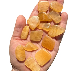 May include: A collection of tumbled yellow calcite stones. The stones are smooth and polished, with a light yellow color. They are arranged in a hand, with the fingers spread out.