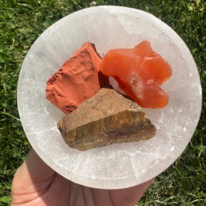 May include: Three tumbled stones in a white selenite bowl. The stones are red jasper, tiger's eye, and carnelian.