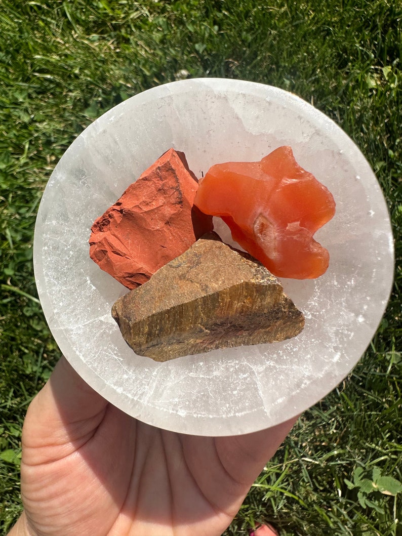 May include: Three tumbled stones in a white selenite bowl. The stones are red jasper, tiger's eye, and carnelian.