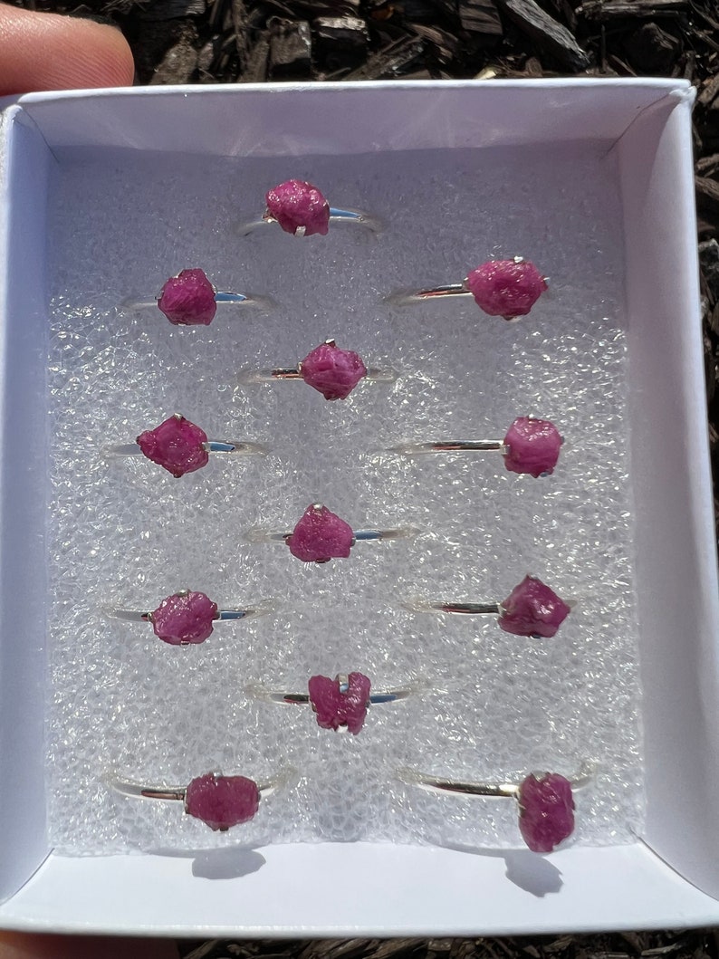 May include: A set of ten silver rings with pink ruby stones. The rings are displayed in a white box with a white foam lining.